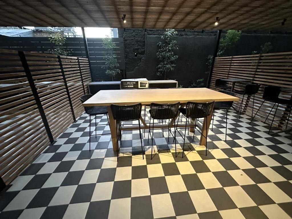 Um chão xadrez, uma mesa com cadeiras e vista para árvores através de uma cerca.