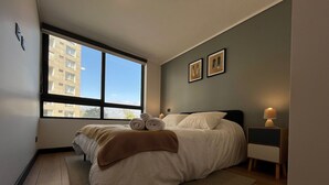 A bedroom with a large window, a bed with rolled towels, a bedside lamp, and two framed pictures on the wall. - Oceana Suites San Eugenio (Santiago)