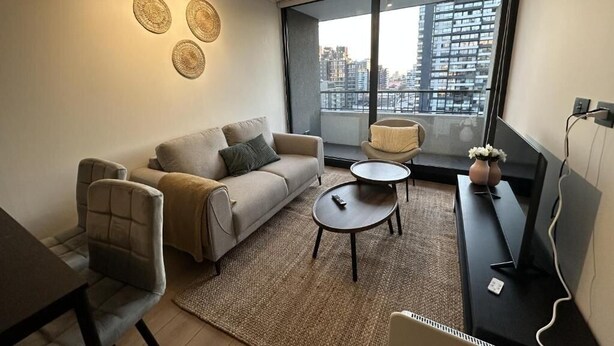 A modern living room with a sofa, armchairs, a coffee table, and a TV. Large windows offer a view of the cityscape. - Oceana Suites San Eugenio (Santiago)