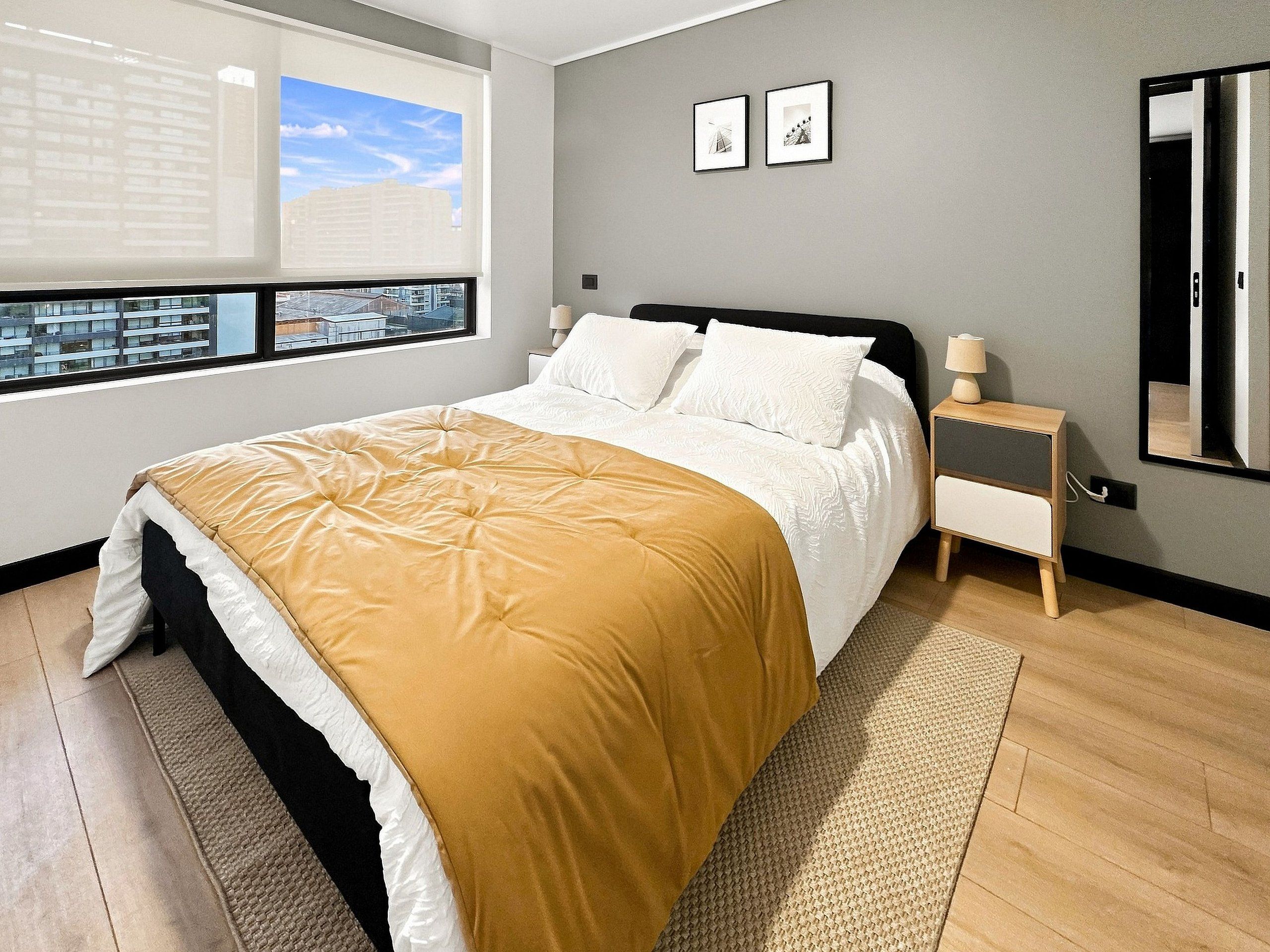 A modern bedroom with a large bed, a bedside table, a mirror, and a window with a city view.