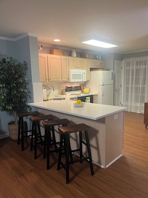 A kitchen with a central island, bar stools, a microwave, a refrigerator, and wooden flooring. - The LemonDrop: 3-bedroom townhome in charming Lewes minutes away from Beaches! (Lewes)
