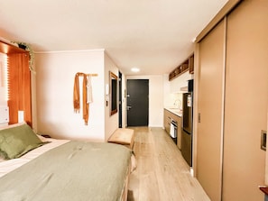 A compact hotel room with a bed, a bench, a small kitchenette, and a door leading to another room. - Downtown Studio - Home Office & Fast WiFi (Santiago)