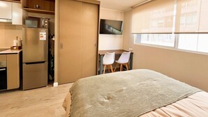 Ein kleines Zimmer mit Bett, Küchenzeile und einem an der Wand befestigten Fernseher.
