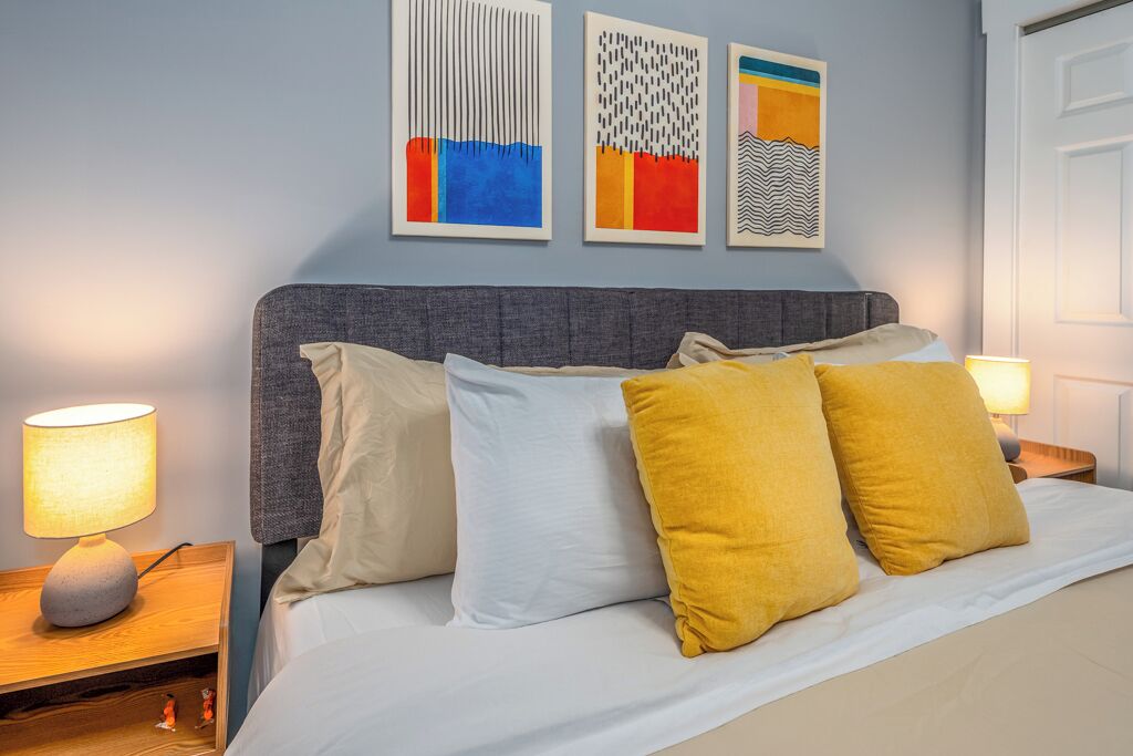Een opgemaakt bed met witte en mosterdkleurige kussens, een grijs hoofdeinde en een nachtkastje met een lamp.