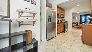 Una cocina moderna con un refrigerador de acero inoxidable, armarios de madera y piso de baldosas. Se observan estantes empotrados y un comedor al fondo.