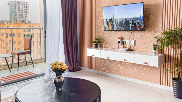 Een moderne woonkamer met een grote televisie aan een wand met een marmerpatroon, een ronde zwarte salontafel, een witte console met een plant, en uitzicht op een stadsgezicht door een balkon.