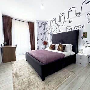 Une chambre moderne avec un lit noir, des rideaux blancs et bruns, un bureau en bois et un mur orné d’œuvres d’art abstraites.