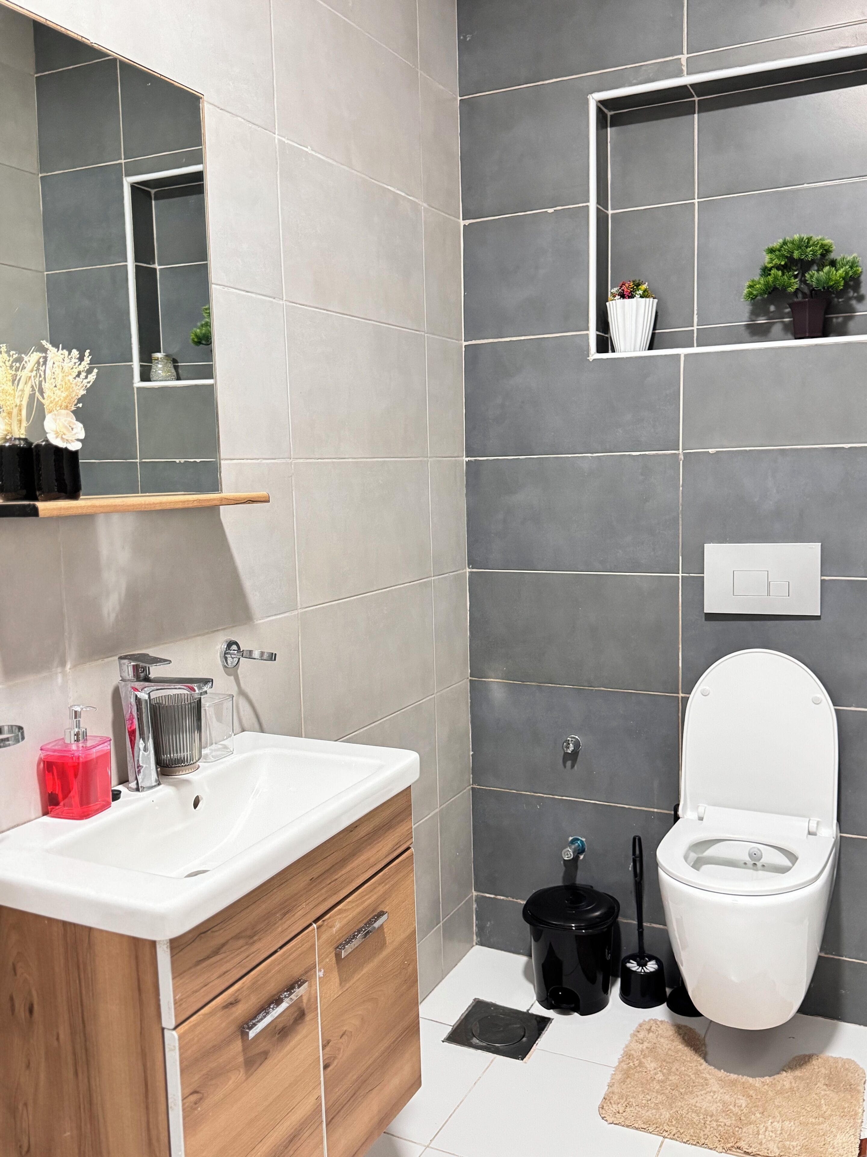 A modern bathroom with a white sink, wooden cabinet, and a toilet. There is a mirror above the sink and a shelf with plants.