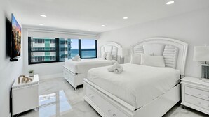 Ein modernes Schlafzimmer mit zwei weißen Betten, einem großen Fenster mit Meerblick und einem an der Wand befestigten Flachbildfernseher.