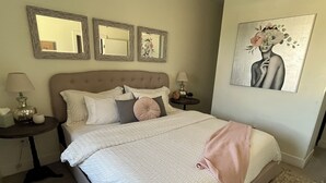 A neatly made bed with a quilted white comforter and a pink throw blanket. There are three bedside tables with lamps and decorative items. The room has a beige carpet and a large painting on the wall. - Downtown rooftop, hot tub retreat adjacent to RiNo & Lodo Walk score of 95! (Denver)