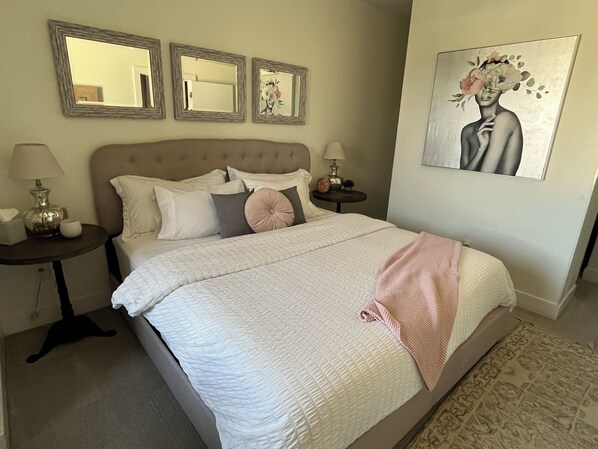 Un lit bien fait, recouvert d’une couette blanche matelassée et d’un plaid rose. Il y a trois tables de chevet avec des lampes et des objets décoratifs. La pièce est dotée d’un tapis beige et d’un grand tableau au mur.