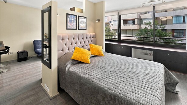 Une chambre moderne avec une tête de lit capitonnée, un grand lit recouvert d’une couette grise texturée, deux coussins jaune vif et une vue sur la ville par une grande fenêtre.
