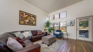 A living room with a brown leather sofa, a grey ottoman, a blue armchair, and a large window with a view of trees. - East / Sabino / 2 BR 2BA / Golf View / Htd Pools (Tucson)