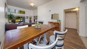 A spacious dining area with a wooden table and white chairs, a seating area with a brown sofa, and a view of the outdoors through large windows. - East / Sabino / 2 BR 2BA / Golf View / Htd Pools (Tucson)