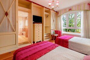 A bedroom with two beds, a TV, a wardrobe, and a window with a view of greenery.
