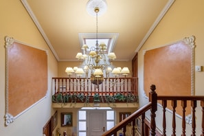 A grand staircase with a chandelier, a balcony with greenery, and a door at the bottom.