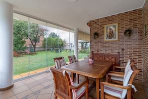 A patio with a wooden table and chairs, a brick wall with a religious painting, and a glass door leading to a grassy outdoor area.