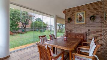 A patio with a wooden table and chairs, a brick wall with a religious painting, and a glass door leading to a grassy outdoor area.