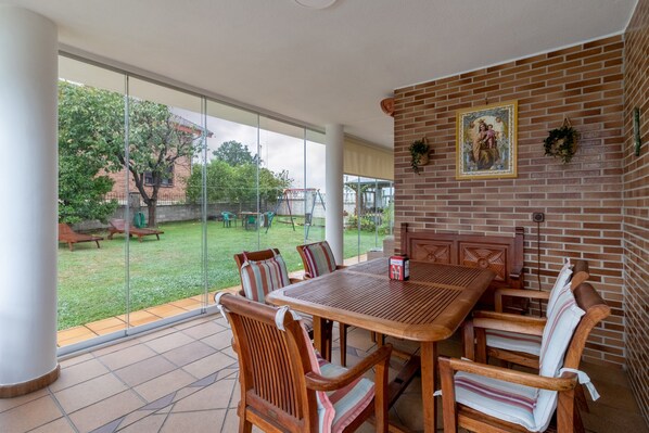 A patio with a wooden table and chairs, a brick wall with a religious painting, and a glass door leading to a grassy outdoor area.