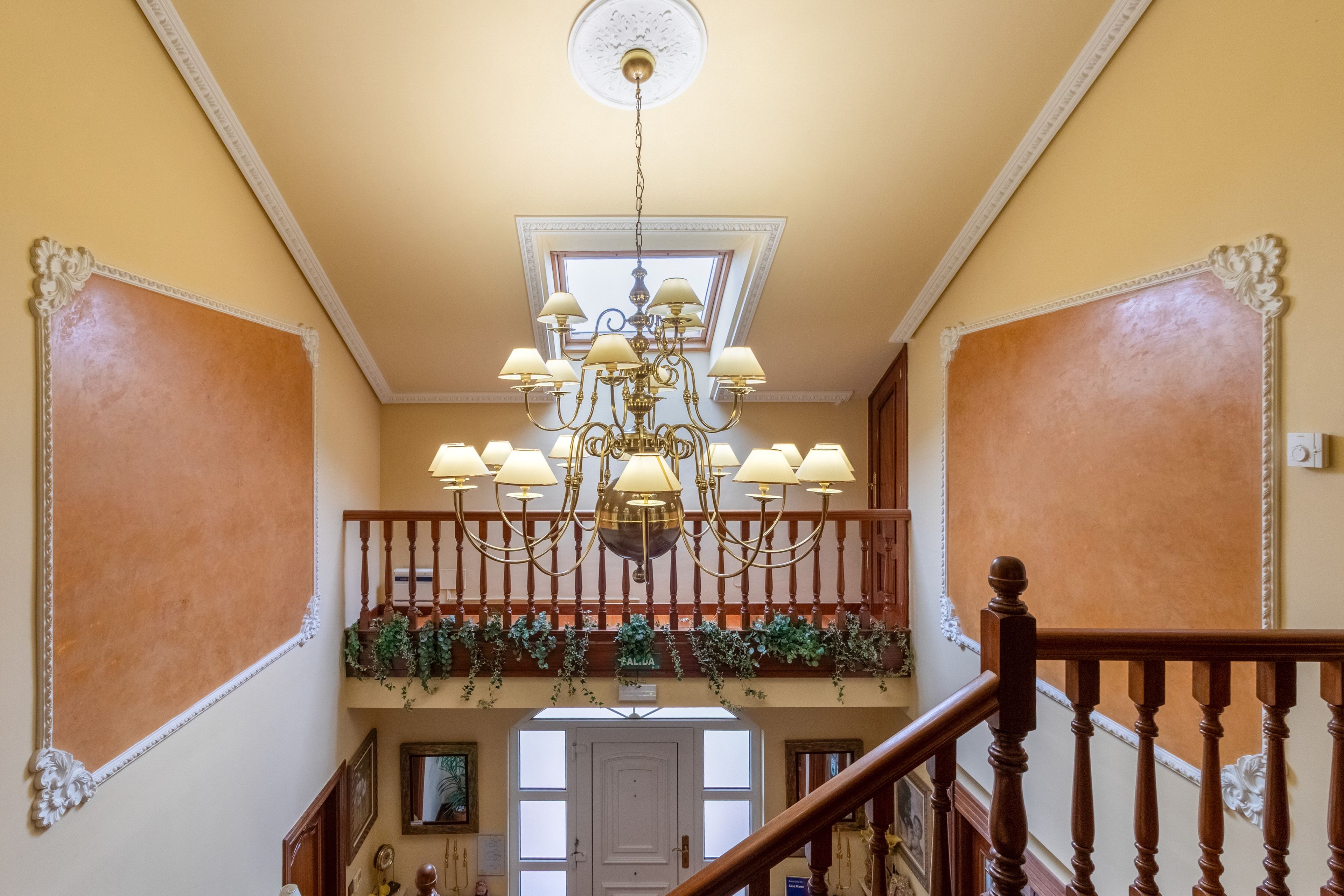 Uma escadaria imponente com um candelabro, uma varanda com vegetação e uma porta na base.