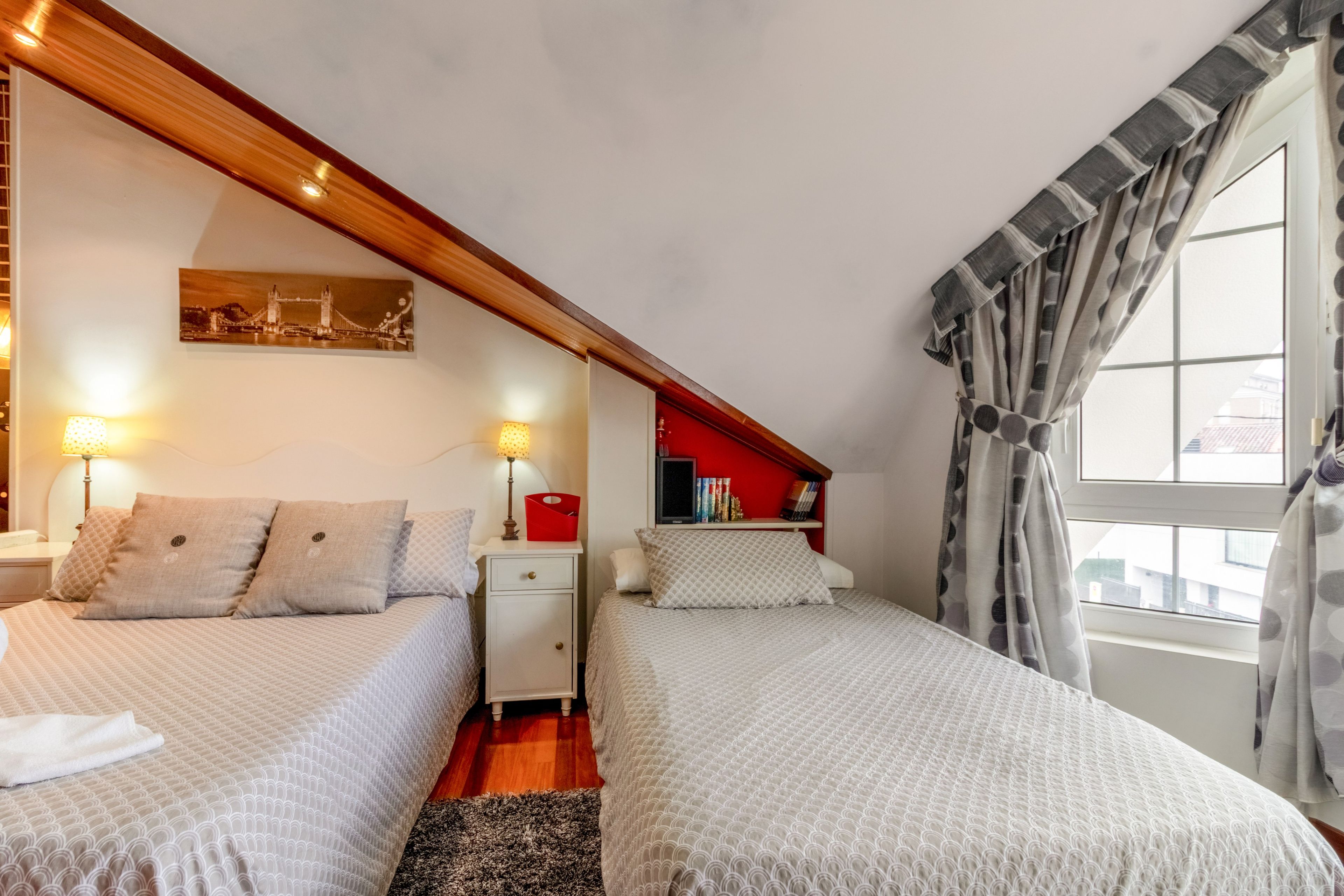 Un dormitorio con dos camas, una mesita de noche, una pared roja, una estantería y una ventana con cortinas.