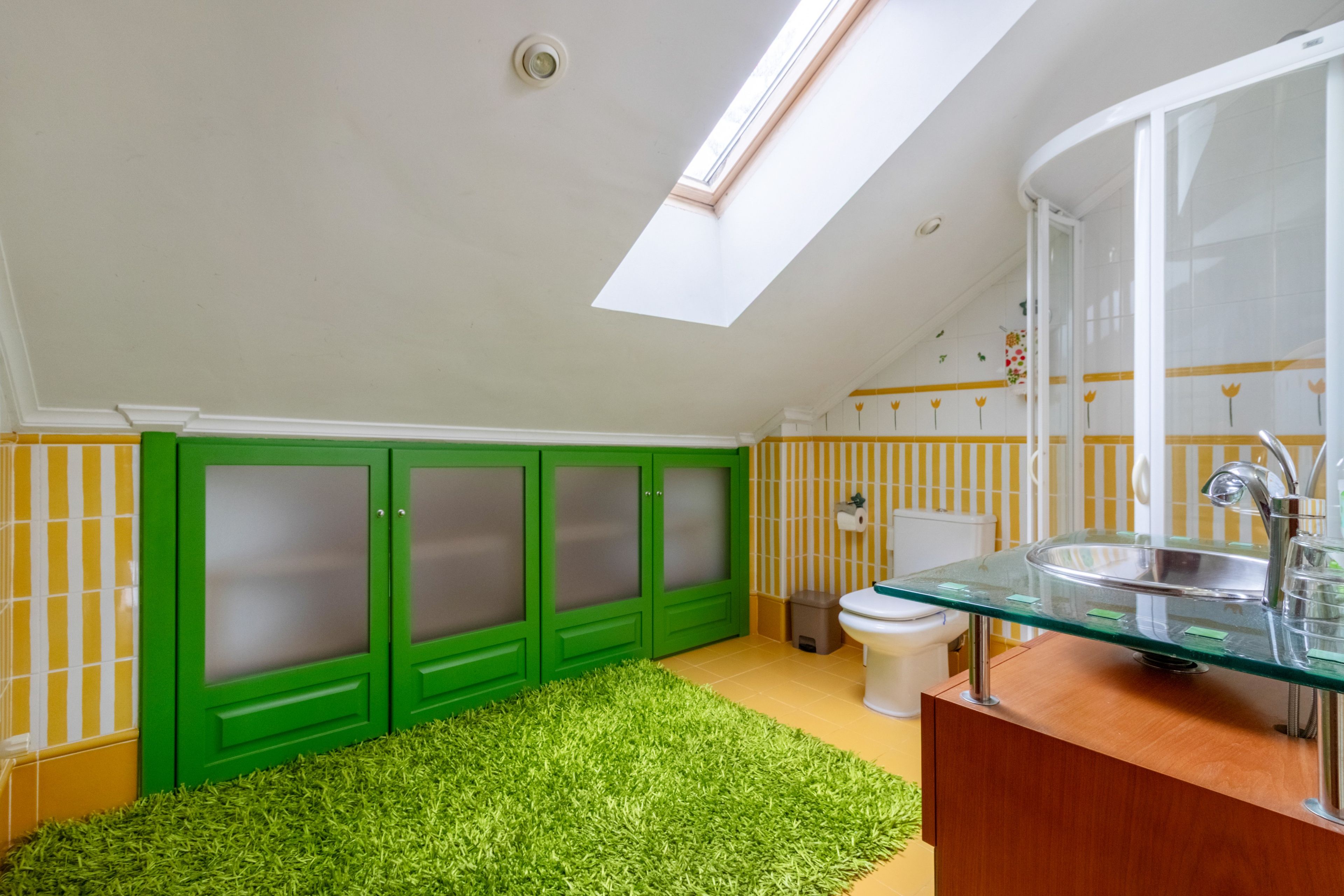 A bathroom with a skylight, green cabinets, a glass sink, and a yellow-striped wall.