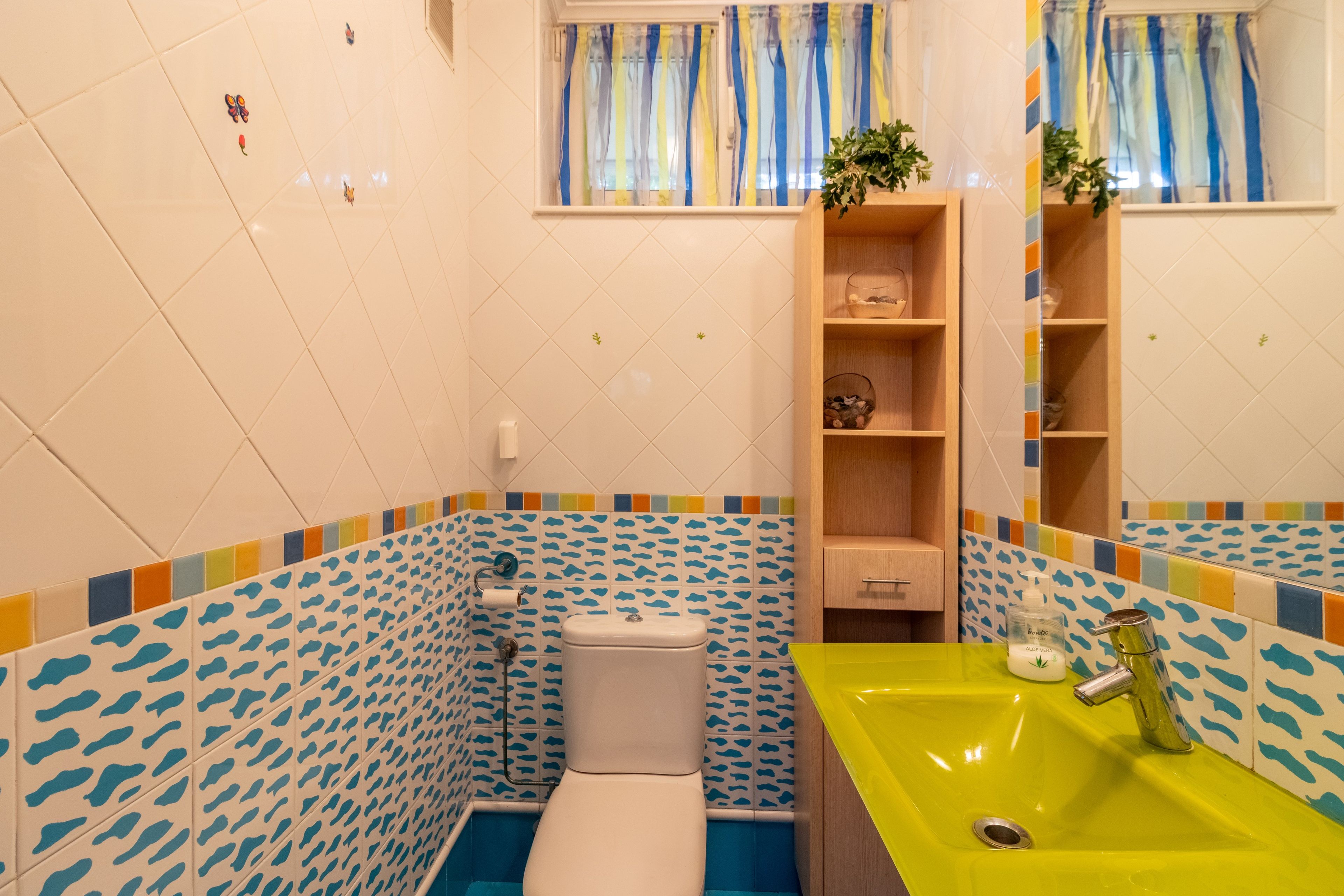 A bathroom with a toilet, a sink, and a shower. There is a shelf with decorative items and a mirror.