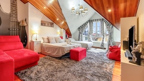 Une chambre avec un plafond en pente, un lit, un canapé rouge, un pouf rouge, une petite table et un miroir.