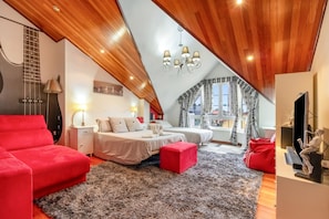 Une chambre avec un plafond en pente, deux lits, un canapé rouge, un pouf rouge, un lustre et une grande fenêtre avec des rideaux.