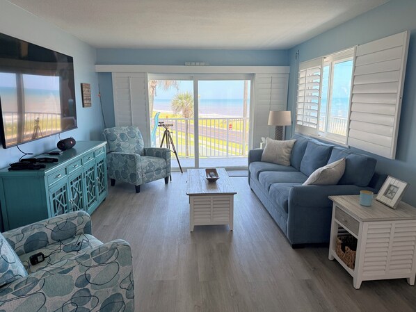 A living room with a TV, a sofa, a chair, and a balcony with a view of the ocean. - Beautiful Beach Condo in Flagler Beach (Flagler Beach)