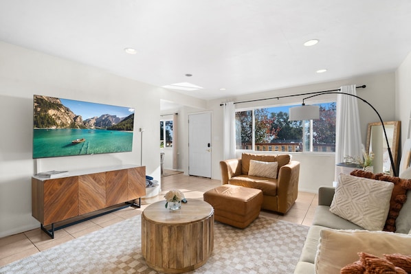 Un salón moderno con un televisor de pantalla plana grande, una mesa de centro de madera, un sofá color beige y vistas a un paisaje montañoso a través de amplias ventanas.