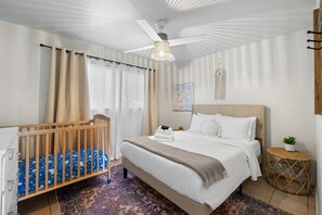 Une chambre avec un grand lit, un berceau en bois, un ventilateur de plafond et une petite table ronde agrémentée d’une plante.
