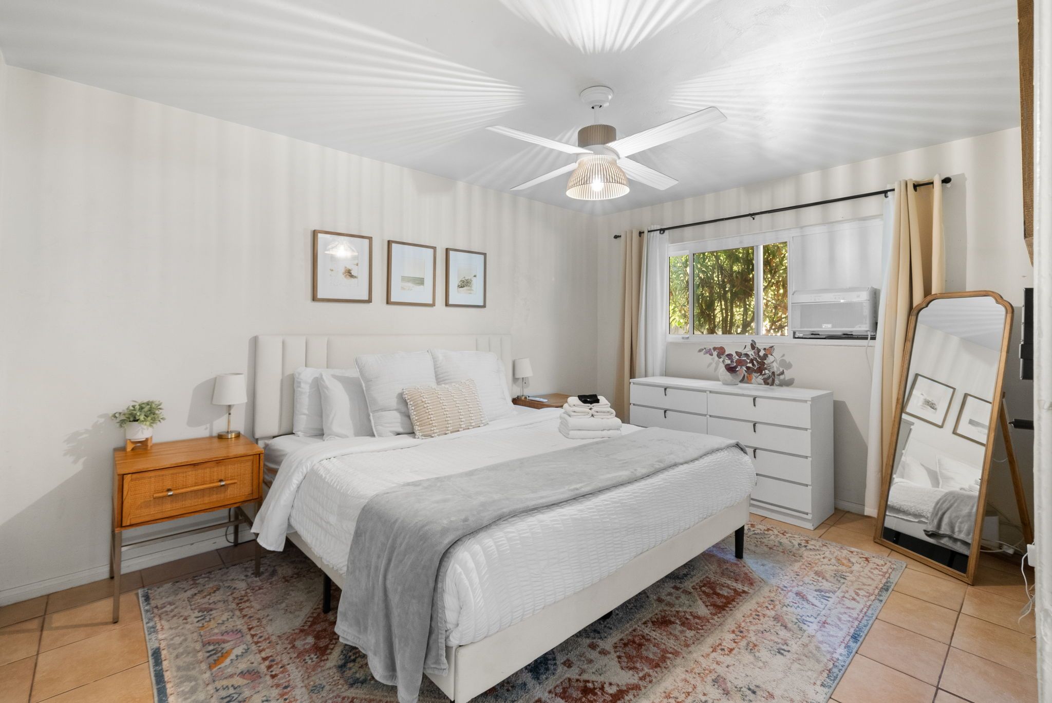 Un dormitorio con cama, mesitas de noche, cómoda, ventilador de techo y ventana con cortinas.