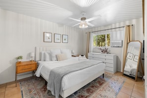 Un dormitorio con cama, mesitas de noche, cómoda, ventilador de techo y ventana con cortinas.