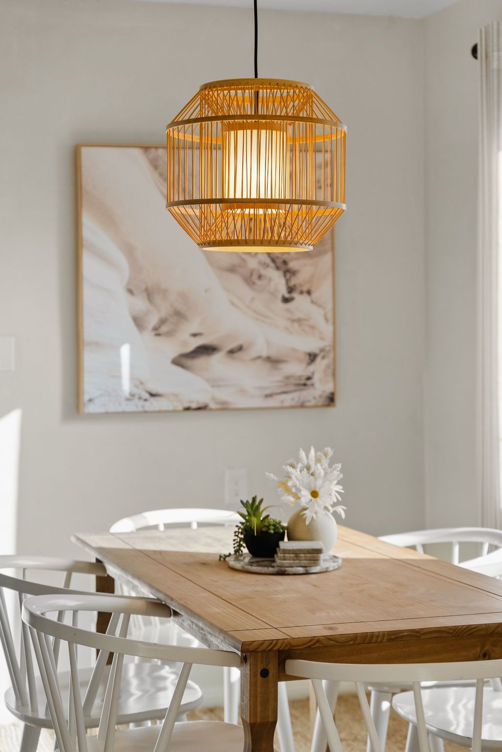 Un comedor con una mesa de madera, sillas blancas, una lámpara colgante y una obra de arte de mármol en la pared.