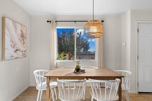 Un coin repas avec une table en bois et des chaises blanches, un luminaire suspendu et une fenêtre donnant sur des arbres et des bâtiments.
