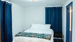 A single bed with white bedding and a floral-patterned throw, flanked by blue curtains, in a room with white walls and a ceiling light fixture. - Hyland Suite 103, Hyland Hotel (Palmer)