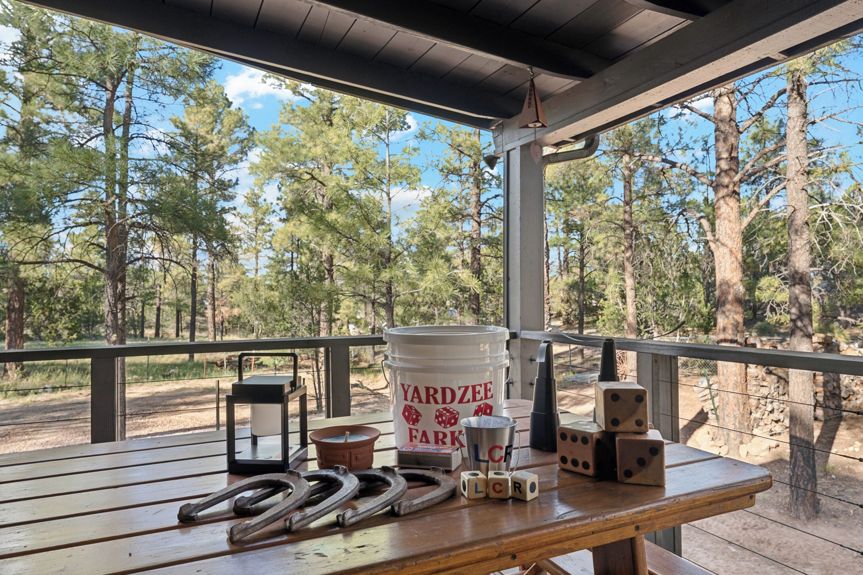 Una mesa de madera con un cubo que dice "Yardzee Farm", dados y herraduras en una terraza con vista a un bosque.