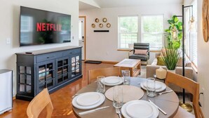 A modern living room with a dining area, a flat-screen TV displaying 'NETFLIX', a dark wooden cabinet, and a large window with blinds. - Quiet/New 2bd Suite w/ Laundry, Kitchen, Wi-Fi (Palmer)