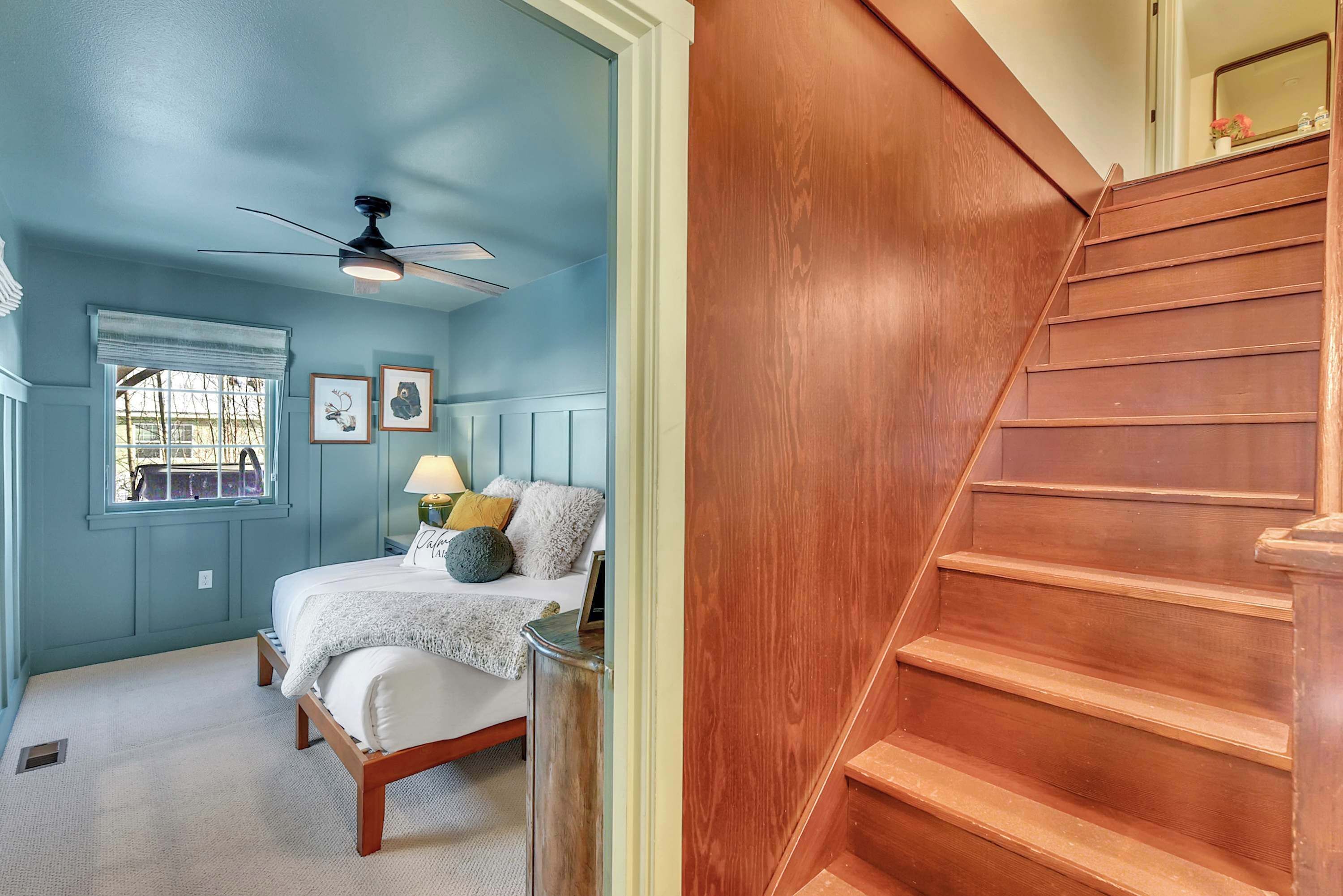 A bedroom with a bed, a ceiling fan, a window, and a wooden staircase.