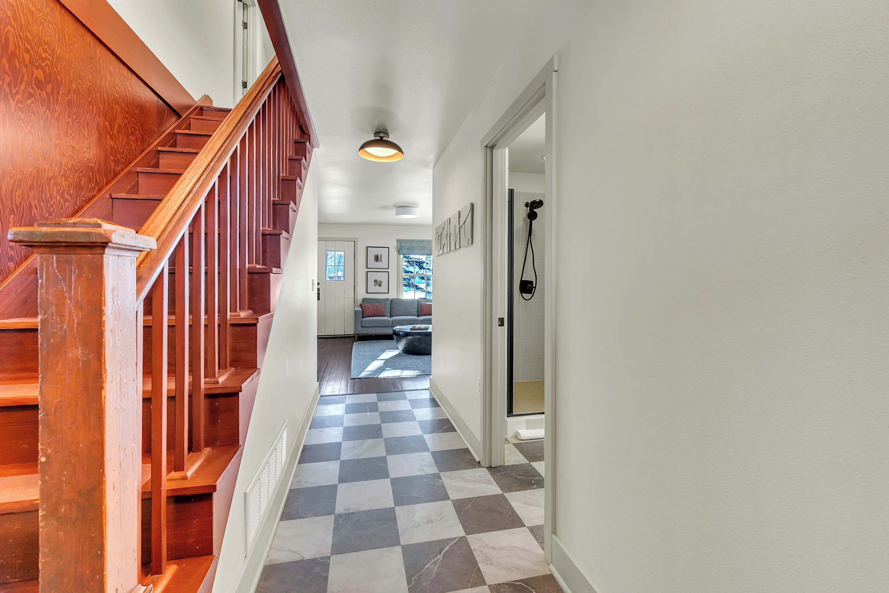 A hallway with a wooden staircase, checkered floor, and a door leading to a living area with a sofa and a coffee table.