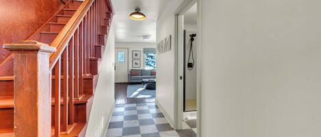 A hallway with a wooden staircase, checkered floor, and a door leading to a living area with a sofa and a coffee table.