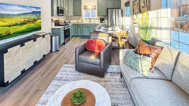 A modern kitchen with a dining area, a sofa, and a large window with a view of a green landscape.