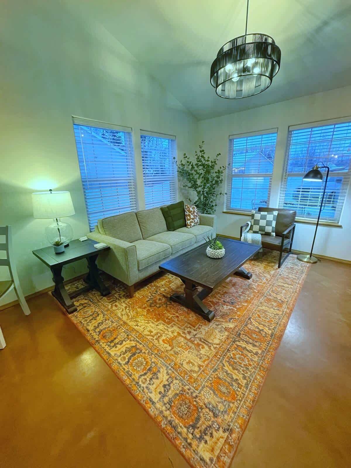 Ein Wohnzimmer mit Sofa, Couchtisch und einer Chandelieber. Es gibt Fenster mit Jalousien, eine Pflanze und einen Teppich.