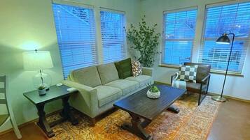 Ein Wohnzimmer mit Sofa, Couchtisch und einer Chandelieber. Es gibt Fenster mit Jalousien, eine Pflanze und einen Teppich.