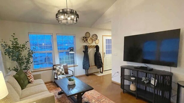 A spacious living room with a large TV, a coffee table, and a sofa. There are windows with blinds, a chandelier, and a rug. - Private & Quiet! 2bd/2ba w/ Laundry & Mt. Views! (Palmer)