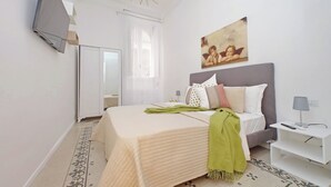 Un dormitorio con cama, mesita de noche, televisor montado en la pared y ventana con cortinas.