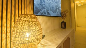 A modern interior with a wooden slat wall, a mounted TV displaying a snowy mountain, and a decorative lamp on a white console. - Seaside, cosy apartment + parking, facing Santa Lucia harbour (Saint-Raphaël)