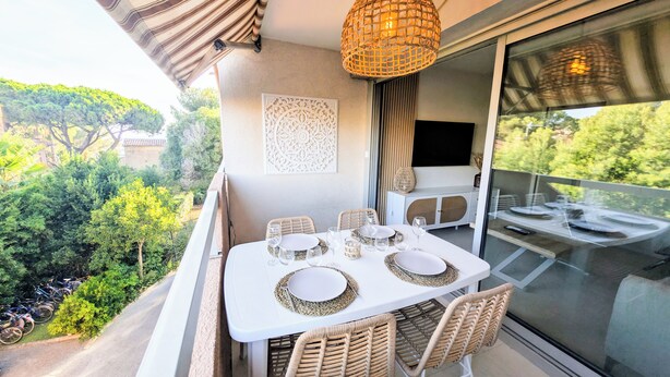 A balcony with a table set for dining, wicker chairs, a decorative wall piece, and a large window with a view of greenery. - Seaside, cosy apartment + parking, facing Santa Lucia harbour (Saint-Raphaël)