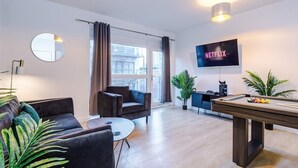 Een moderne woonkamer met een bank, fauteuil, salontafel en een pooltafel. Een televisiescherm toont 'NETFLIX' en er is een raam met gordijnen.